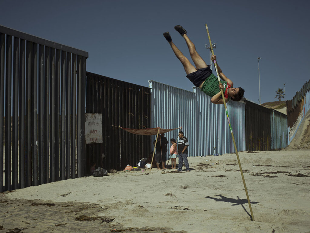 A Mexican pole vault jumper trains by the wall in Tijuana beach while a family uses the wall to mount their beach day tent, Mexico, 2019